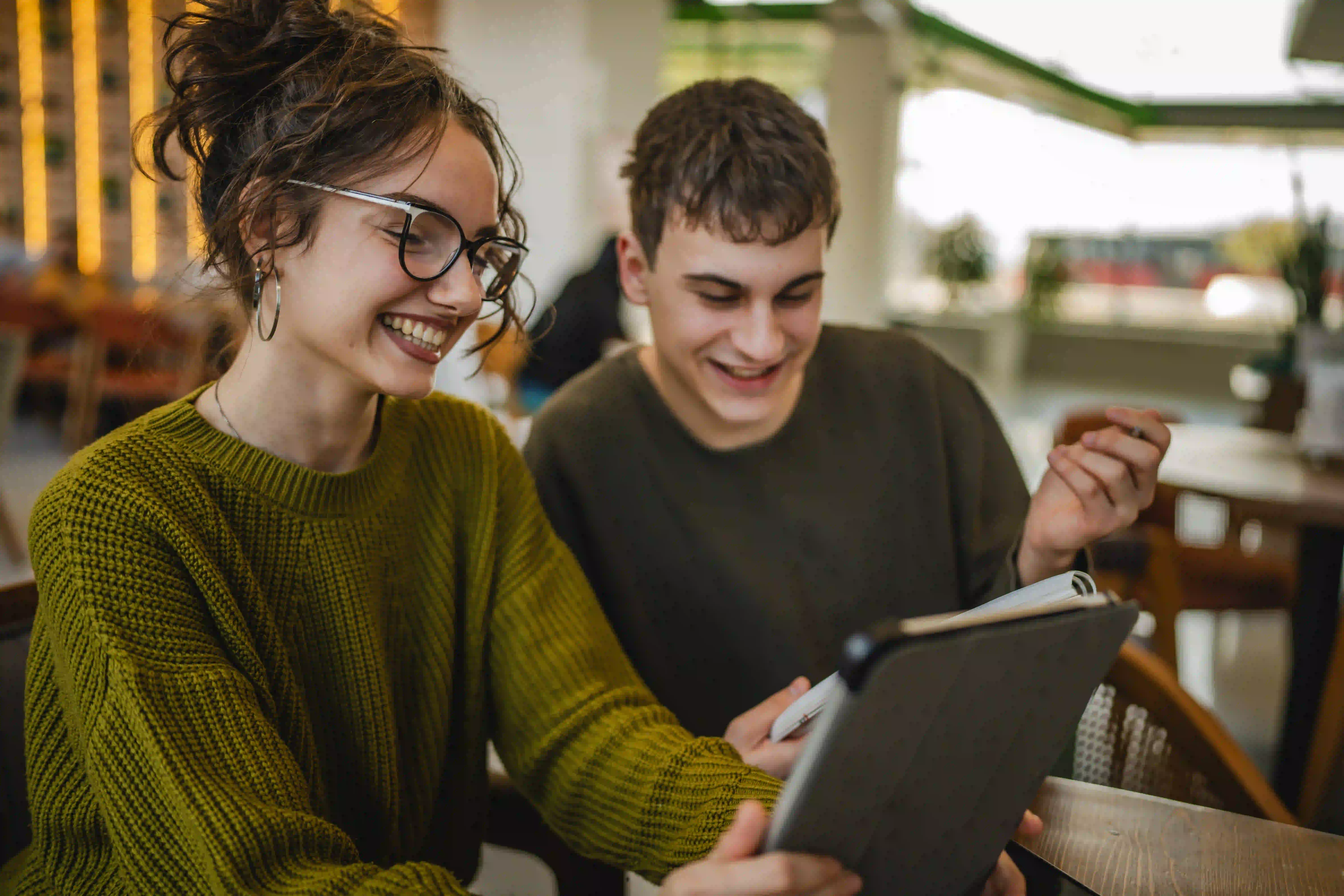 Junge Frau und junger Mann lernen gemeinsam in einem Café und lächeln, während sie ein Tablet betrachten.